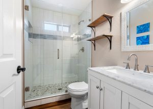 New bathroom addition with glass-enclosed shower, pebble tile floor, white vanity, and open wood shelving