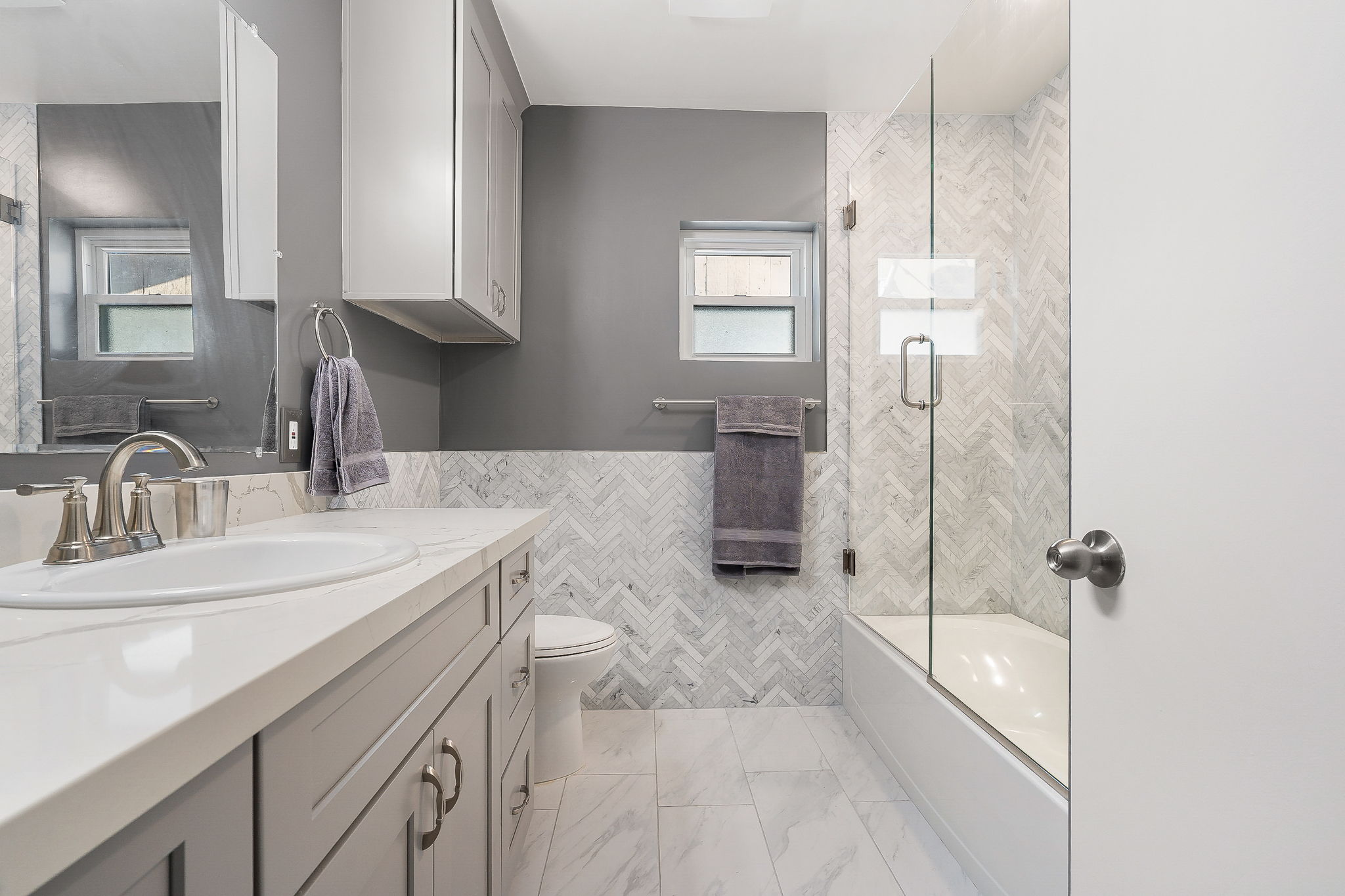 Master bathroom remodel featuring modern shaker cabinets with a white quartz countertop, brushed nickel faucet, and ample storage. The bathroom showcases a large glass-enclosed shower and bath combo with a stylish marble herringbone tile design. Soft gray walls and white ceramic tiled floor complete the look.