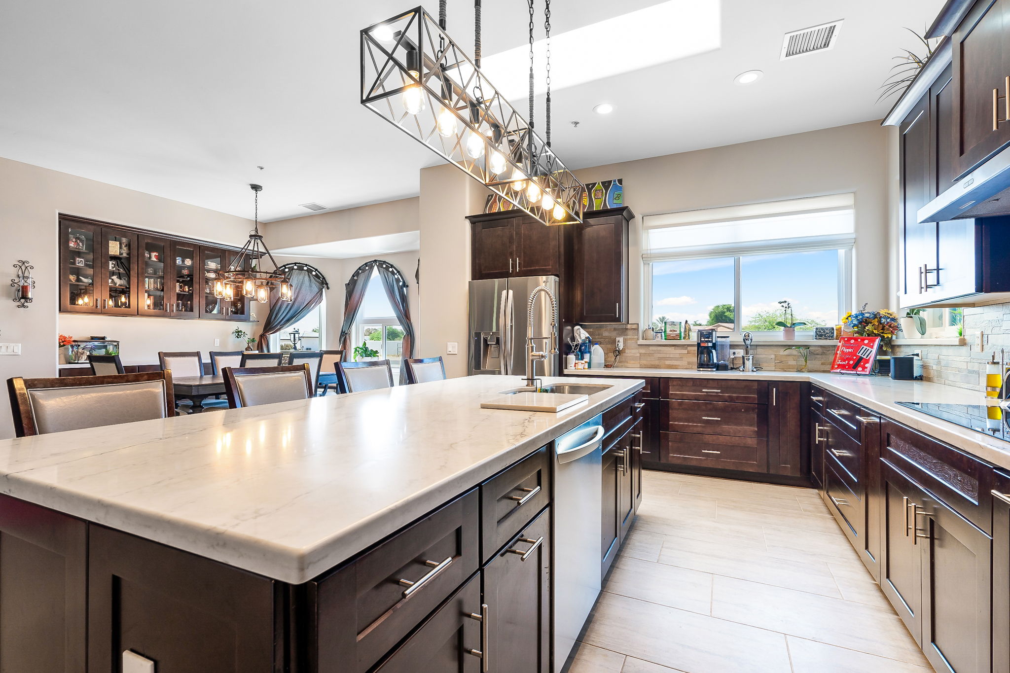 This image showcases a beautifully remodeled kitchen featuring a large, sleek marble island with dark wooden cabinets beneath. Above the island, there is a modern, rectangular chandelier with exposed bulbs, adding an industrial yet elegant touch. The kitchen boasts stainless steel appliances, including a refrigerator and a dishwasher, and has a spacious layout with plenty of counter space. The dining area is visible in the background, with a matching chandelier and dark wood furniture. Large windows allow natural light to flood the space, highlighting the sophisticated and contemporary design.