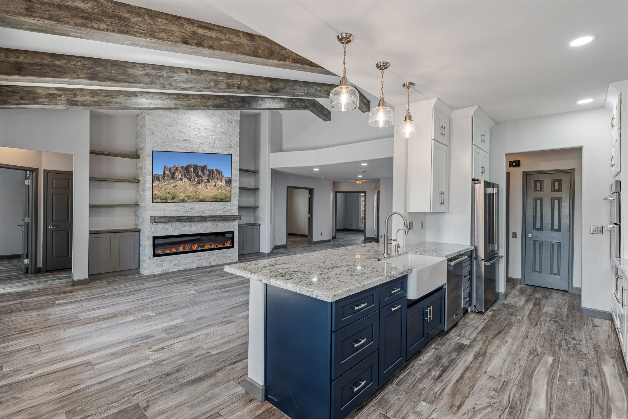 Open-concept living area with exposed wooden beams, an electric feature wall fireplace with a mounted TV, grey cabinetry, and a kitchen featuring a blue island, farmhouse sink, granite countertops, and stainless steel appliances.