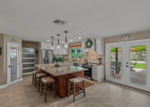 Bright, open kitchen with white shaker cabinets, large granite island, pendant lighting, stainless steel appliances, and French doors leading to a backyard patio.