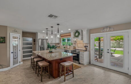Bright, open kitchen with white shaker cabinets, large granite island, pendant lighting, stainless steel appliances, and French doors leading to a backyard patio.