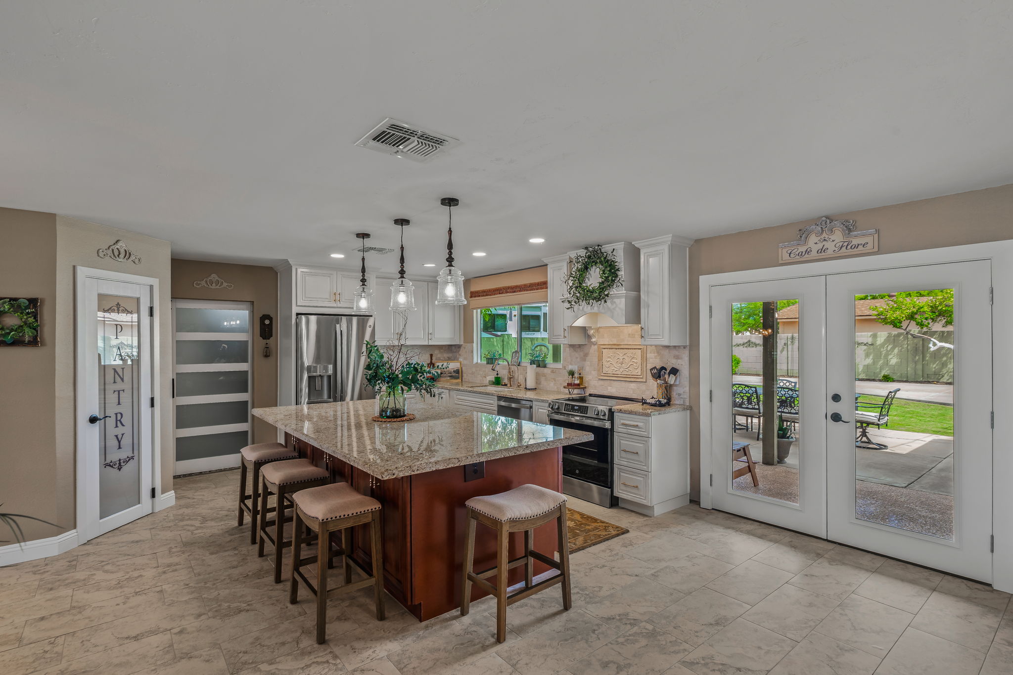 Bright, open kitchen with white shaker cabinets, large granite island, pendant lighting, stainless steel appliances, and French doors leading to a backyard patio.