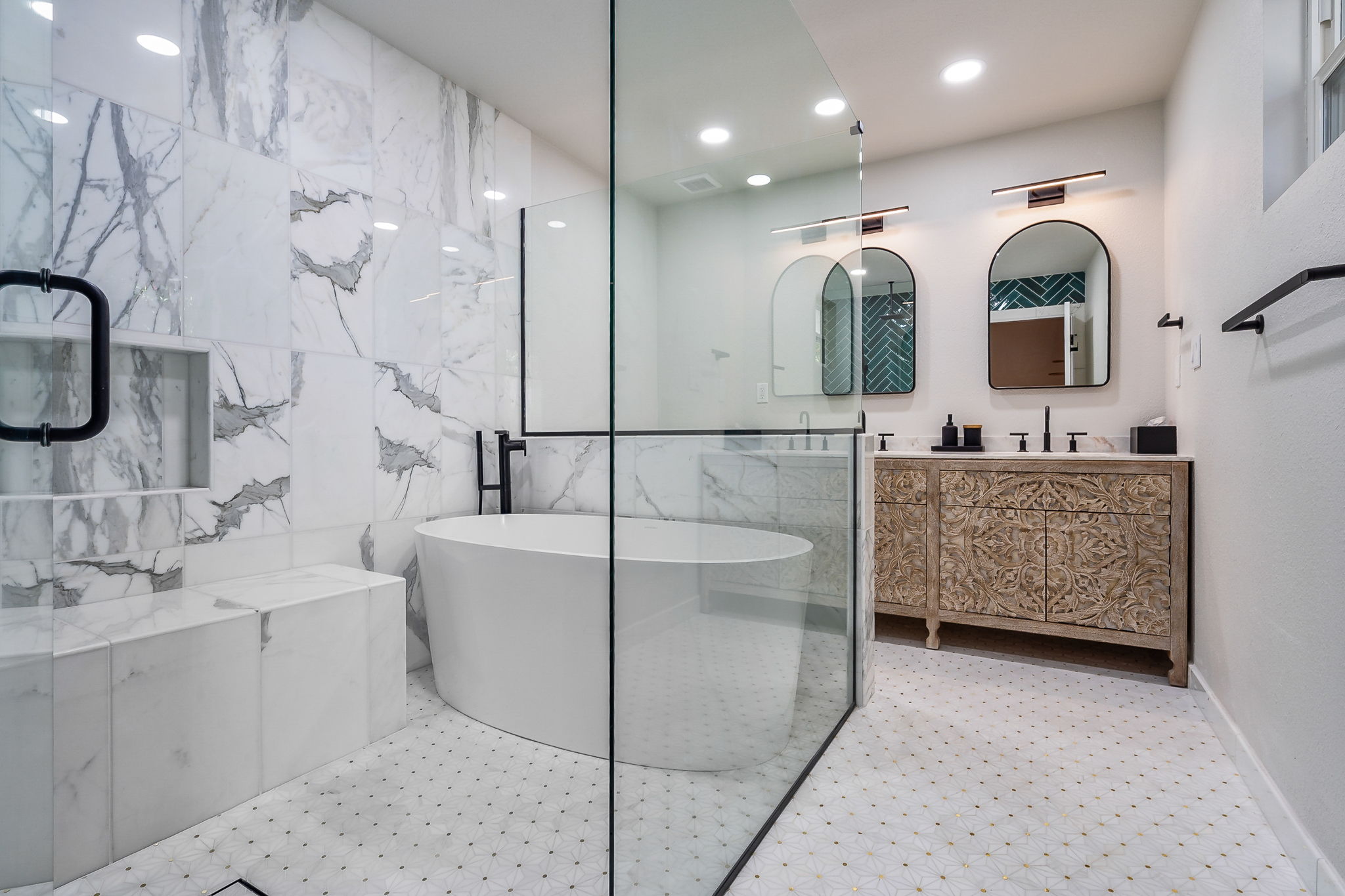 ChatGPT This image shows a luxurious master bathroom featuring a modern, open design. The space is highlighted by a spacious glass-enclosed shower with large, white marble tiles that have striking gray veining. Inside the shower, there is a built-in bench and niche for convenience. A sleek, freestanding bathtub is positioned next to the glass enclosure. The double vanity has intricately carved wooden cabinets, adding a touch of warmth and elegance. Above the vanity are two arched mirrors, each with modern light fixtures. The floor is adorned with a white hexagonal tile pattern with subtle gold accents, completing the sophisticated look of the bathroom.